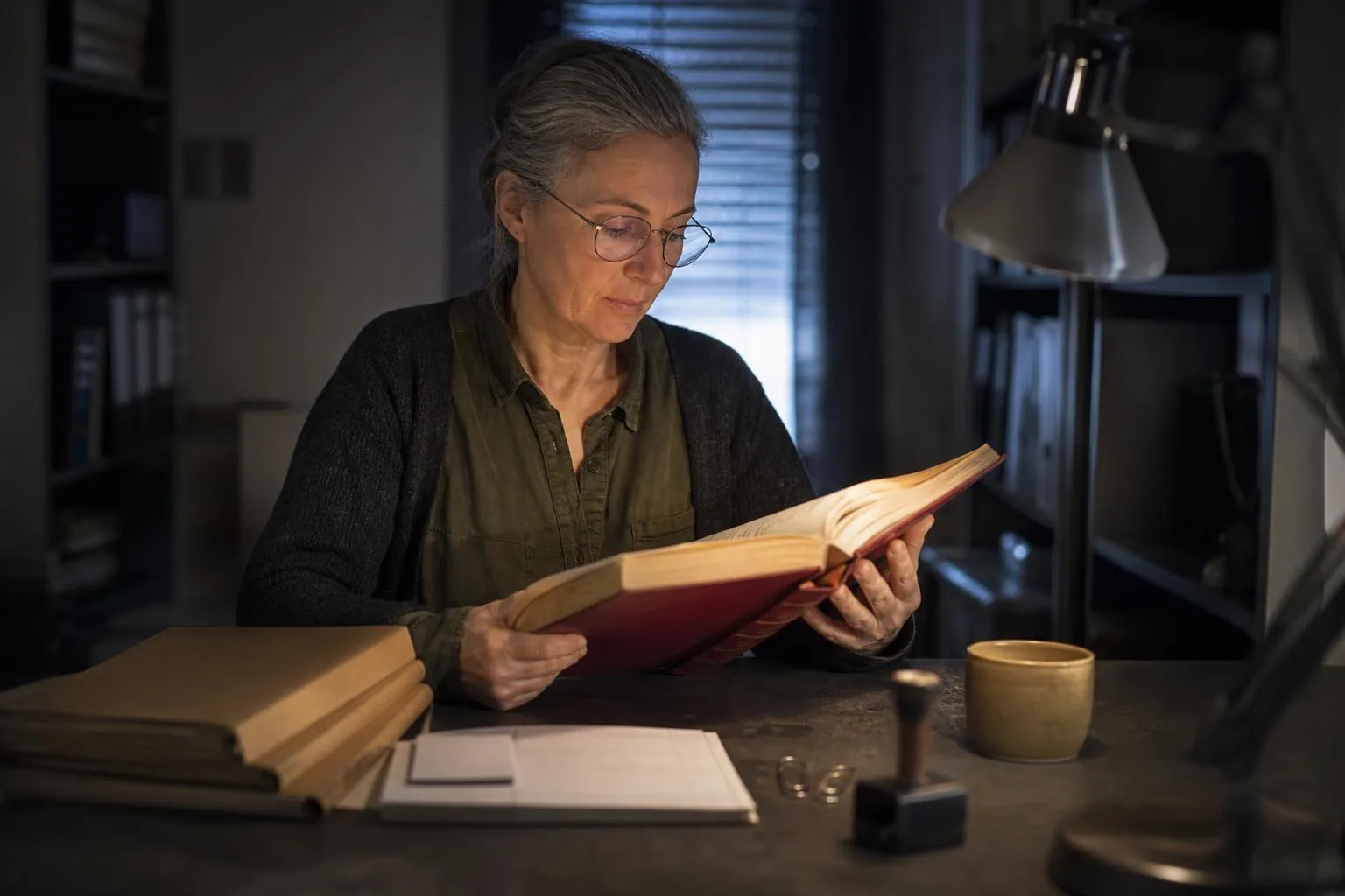 Femme consultant un recueil de textes relié dans un bureau sombre, chemises cartonnées empilées et lampe d’atelier allumée
